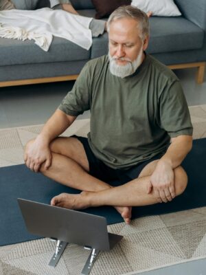 Older man practicing yoga while following an online session on a laptop in his living room.