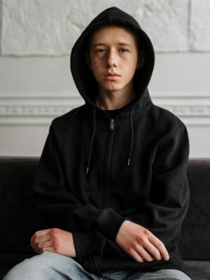 Teenager in black hoodie sitting indoors, contemplating with a serious expression.