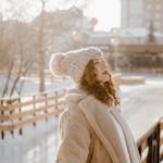 Young woman in winter attire enjoying a sunny day outside, leaning on a wooden railing.